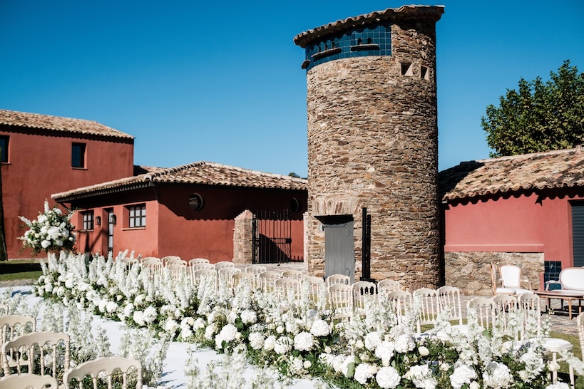 Outdoor wedding ceremony setup with white chairs and floral decorations in front of a rustic building. - ACCAELLE Events | Organizzazione eventi e Wedding planner