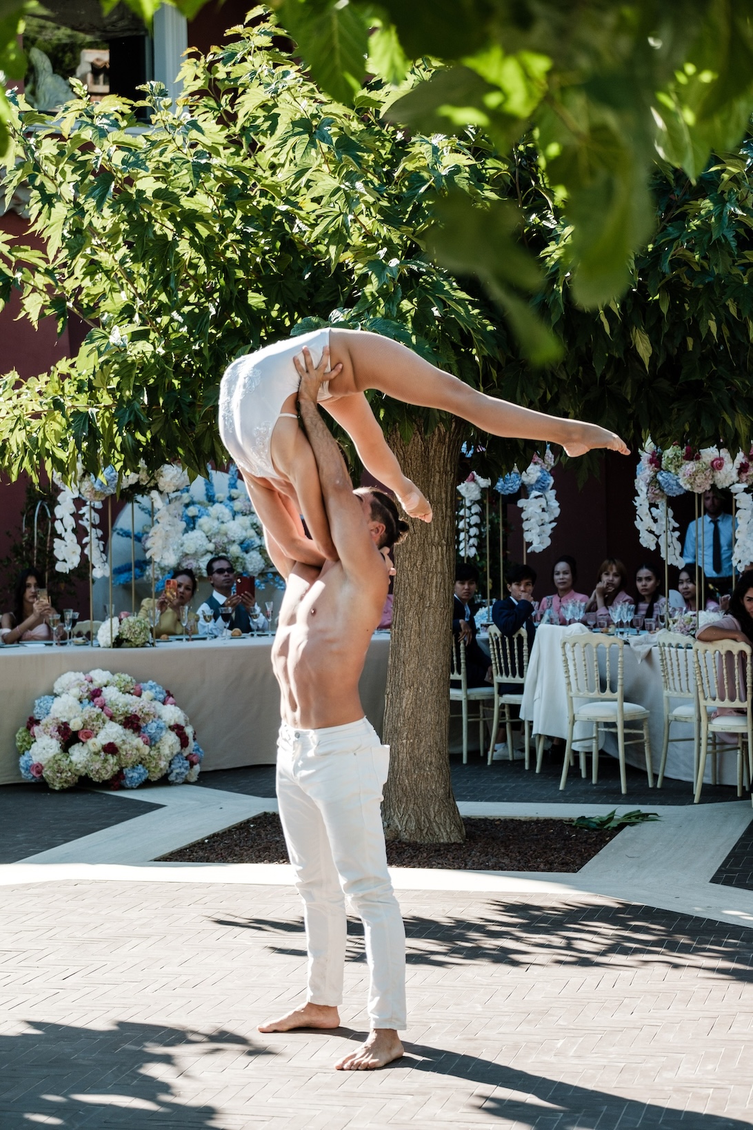 Dancers in white dresses dancing in a courtyard with tree and guests sitting at table in the background - ACCAELLE Events | Organizzazione eventi e Wedding planner