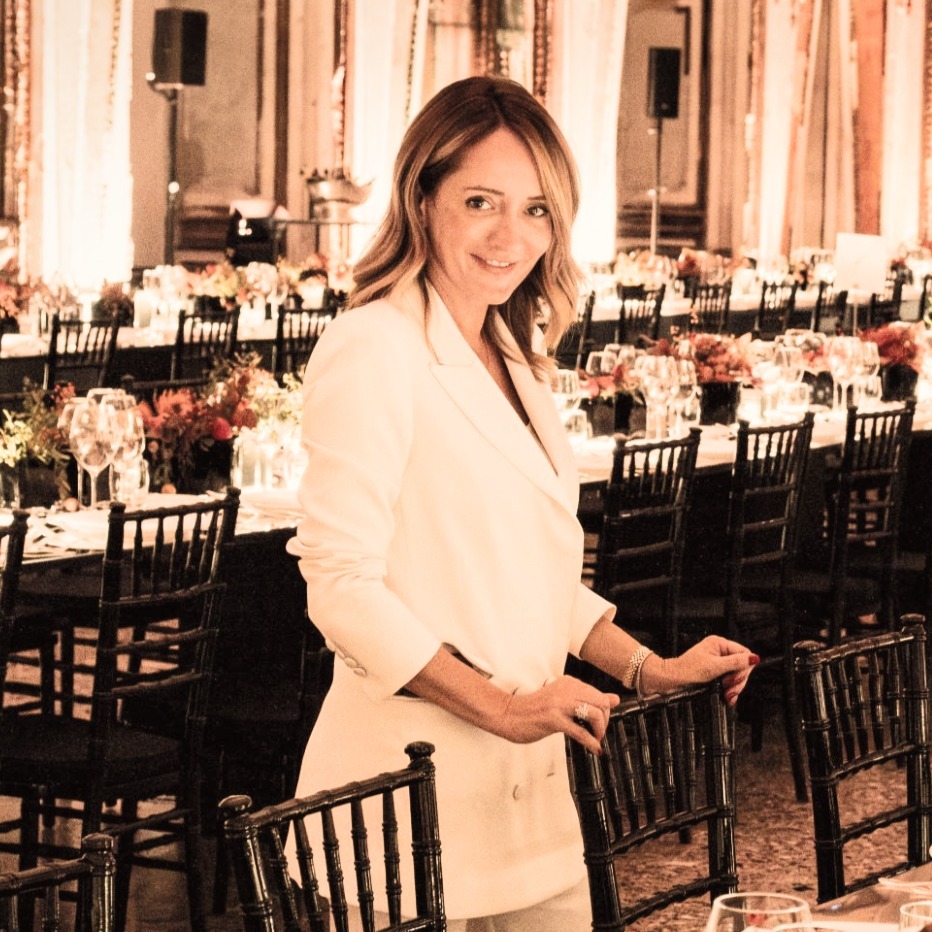 A woman in elegant attire smiles next to a long table set up in a formal room. - ACCAELLE Events | Organizzazione eventi e Wedding planner