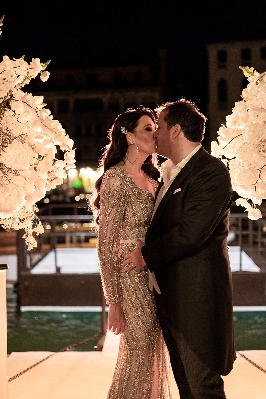 Husband and wife kissing in an evening atmosphere with white flowers behind them - ACCAELLE Events | Organizzazione eventi e Wedding planner