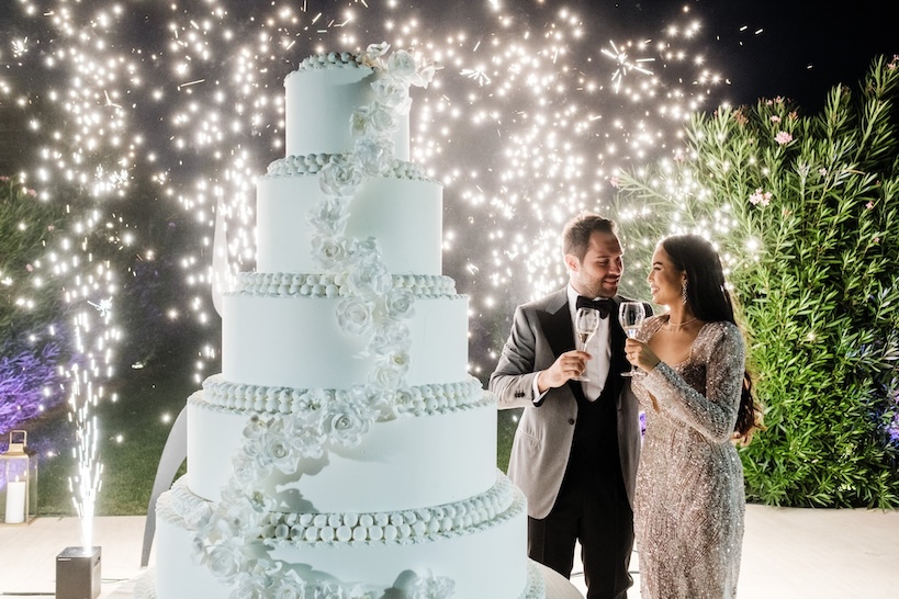 Husband and wife pose with glasses in hand next to wedding cake and white sparkles in the background - ACCAELLE Events | Organizzazione eventi e Wedding planner