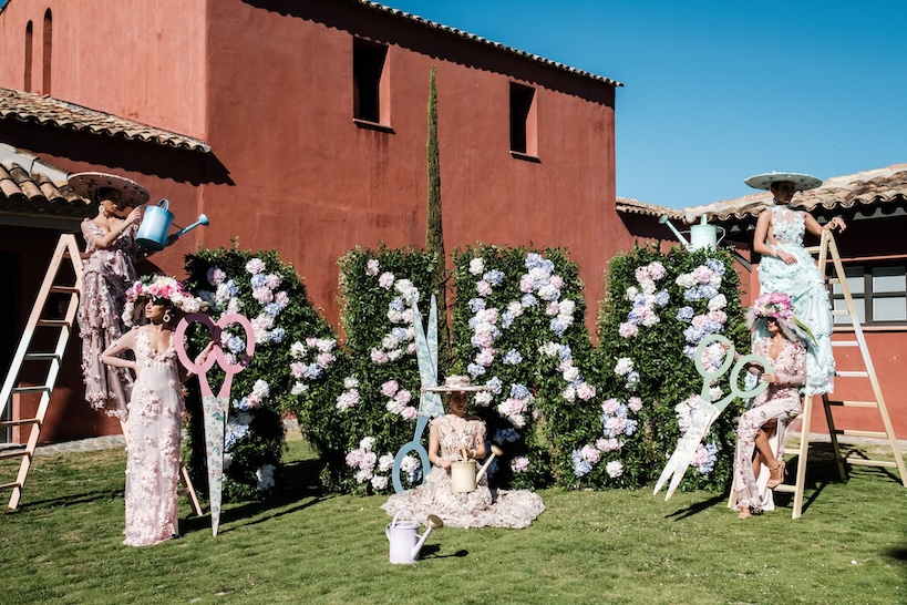 Models in elegant dresses pose in front of large floral letters with giant scissors in a garden with rustic building - ACCAELLE Events | Organizzazione eventi e Wedding planner