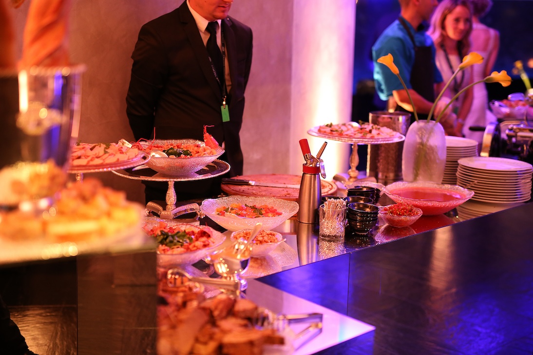 Overview of a rich buffet table in a brightly lit room with guests in the background - ACCAELLE Events | Organizzazione eventi e Wedding planner