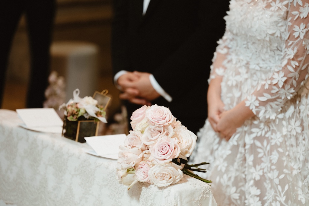 Close-up of the bride and groom's bust and hands with pink flowers and a wedding booklet - ACCAELLE Events | Organizzazione eventi e Wedding planner