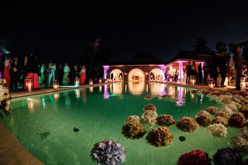 Close-up of a swimming pool with a rustic structure in the background with white, pink and lilac flowers soaking in the eveni - ACCAELLE Events | Organizzazione eventi e Wedding planner