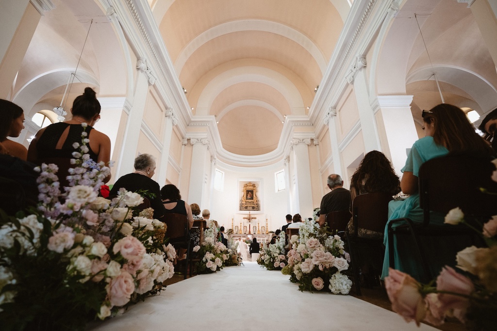Shot from below of the central nave, flowers as ornaments and altar with the bride and groom in the background - ACCAELLE Events | Organizzazione eventi e Wedding planner