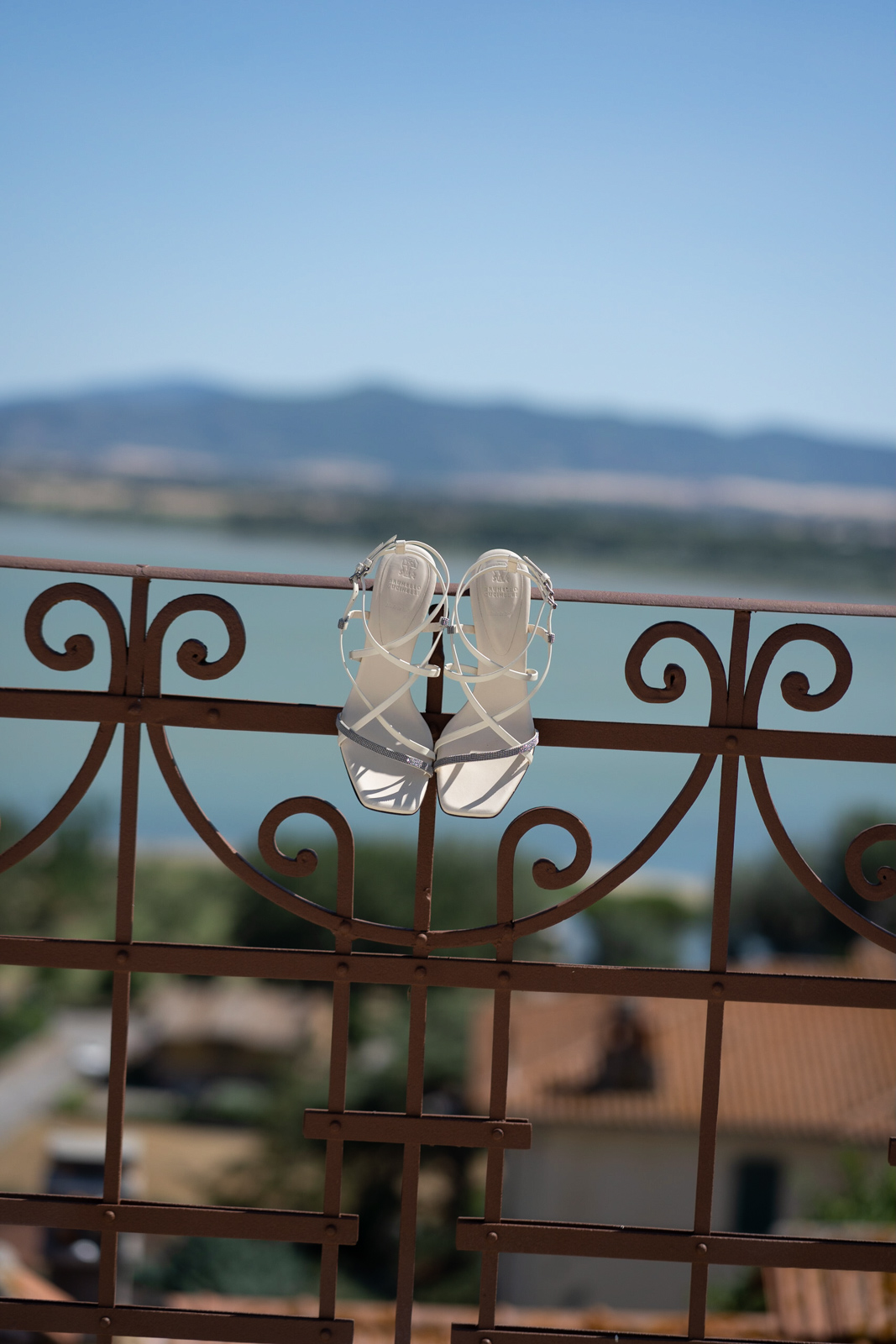 High-heeled wedding shoes hanging from a wrought iron railing with a lake in the background. - ACCAELLE Events | Organizzazione eventi e Wedding planner