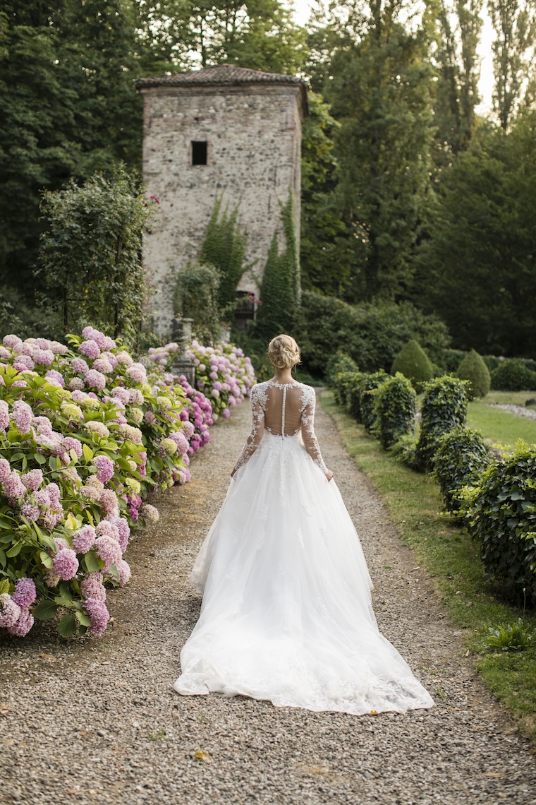 Bride from behind walking towards a ruin in a meadow with hedges and pink flowers - ACCAELLE Events | Organizzazione eventi e Wedding planner