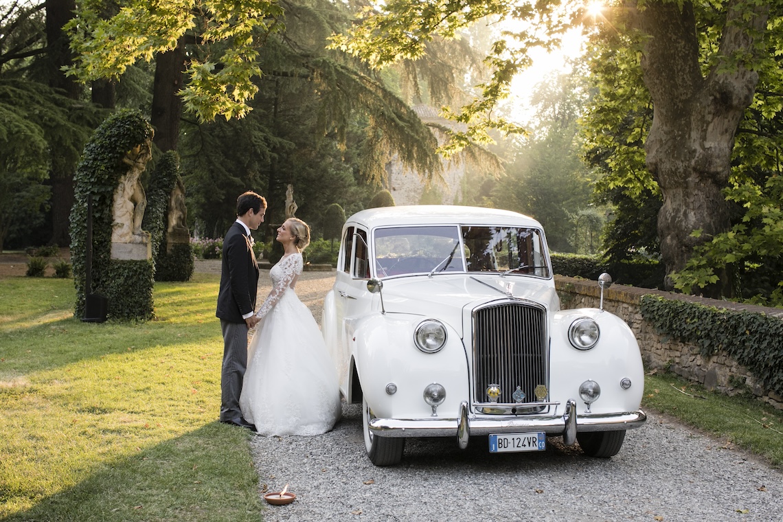 Bride and groom facing each other next to the white wedding car in a garden at sunset - ACCAELLE Events | Organizzazione eventi e Wedding planner