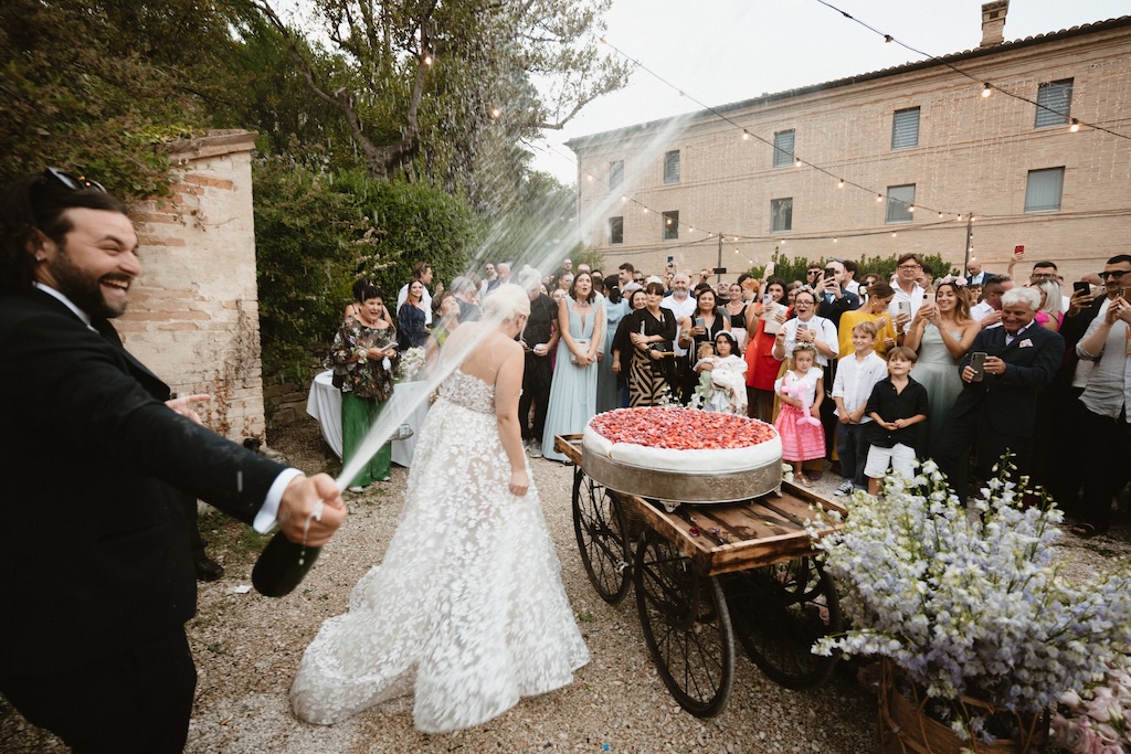 Groom popping champagne with bride, cake and guests in the background - ACCAELLE Events | Organizzazione eventi e Wedding planner