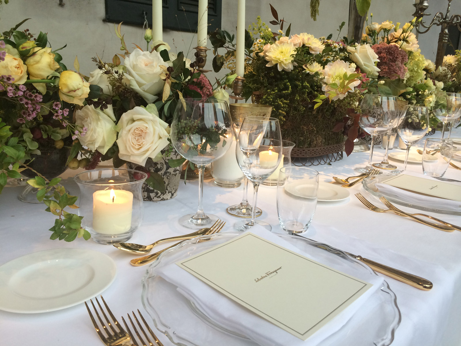 A table set with golden cutlery, a white menu on a plate, and candles among the flowers. - ACCAELLE Events | Organizzazione eventi e Wedding planner
