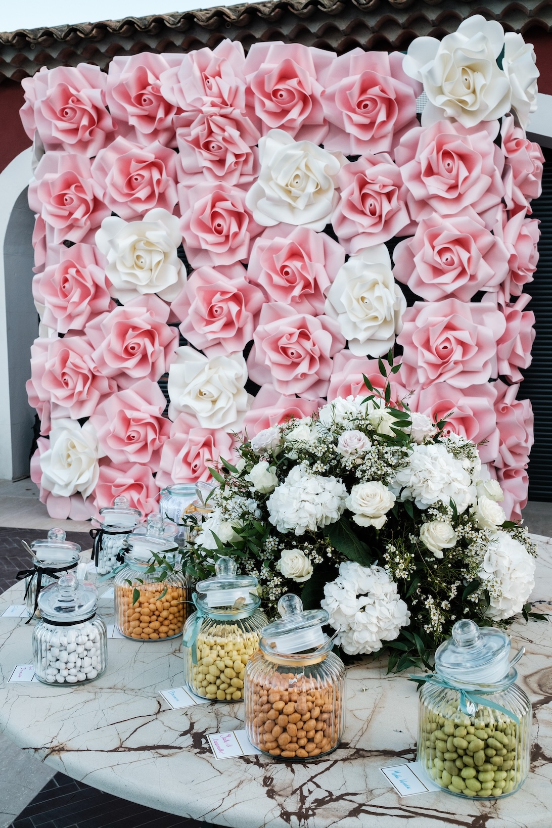 White marble table with coffee tables of various colors and a bouquet of white flowers in the center and a wall of roses in t - ACCAELLE Events | Organizzazione eventi e Wedding planner