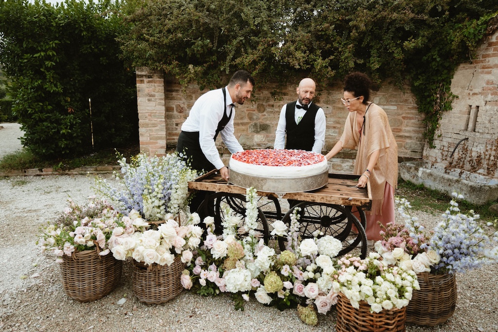 A white and red wedding cake sits on a counter with countless floral bouquets of various colors in front of it. - ACCAELLE Events | Organizzazione eventi e Wedding planner