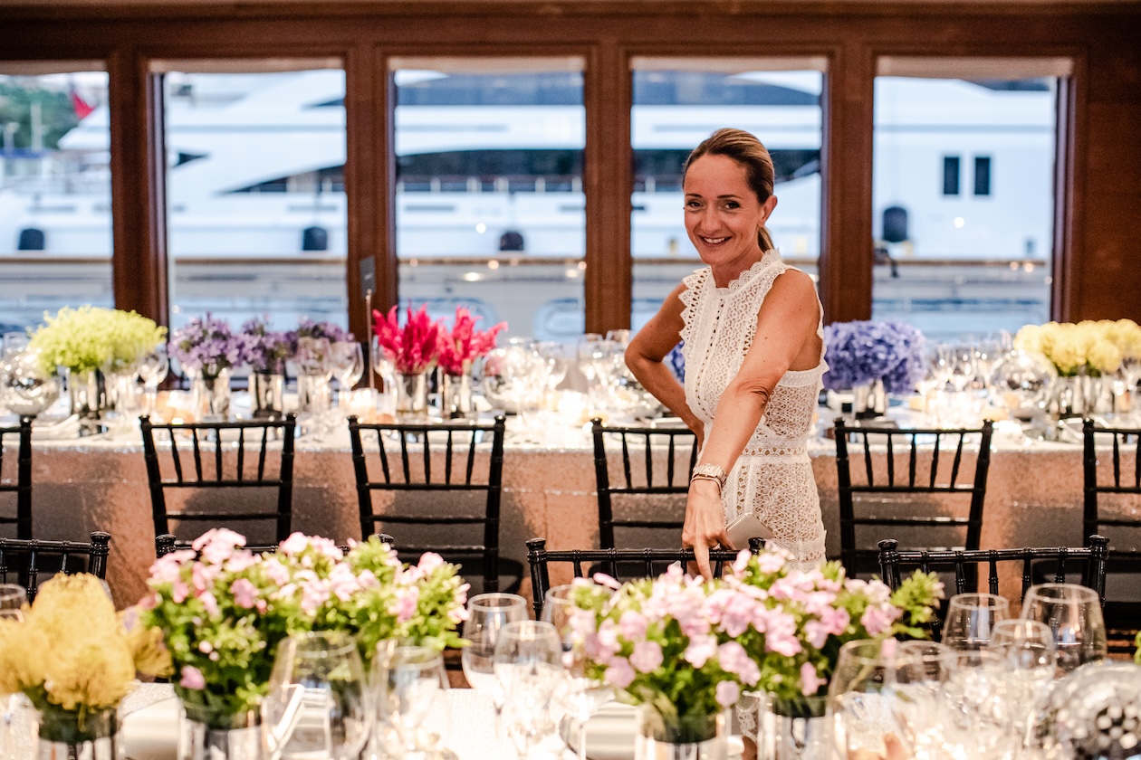 A smiling woman standing behind a flower-filled table inside a yacht. - ACCAELLE Events | Organizzazione eventi e Wedding planner