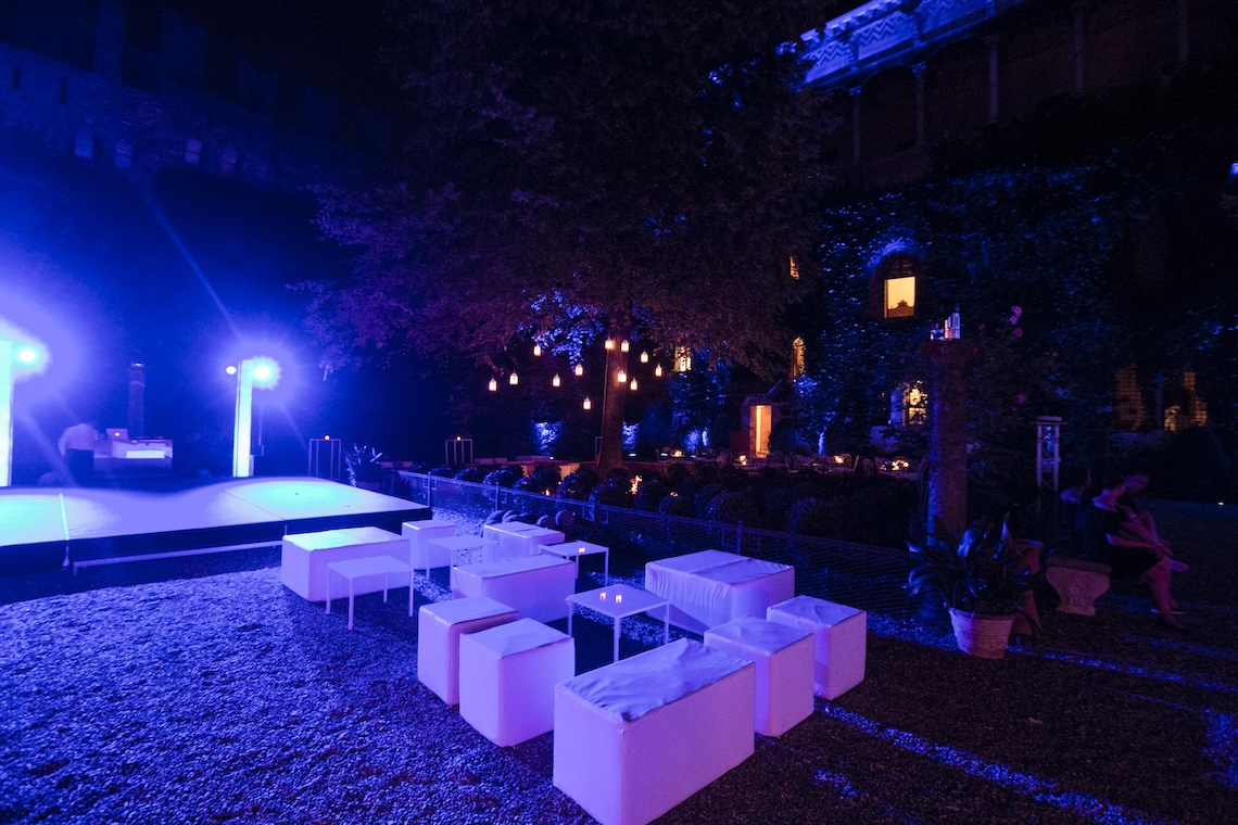 Area lounge esterna serale con pouf bianchi quadrati illuminati da intense luci blu e viola. - ACCAELLE Events | Organizzazione eventi e Wedding planner
