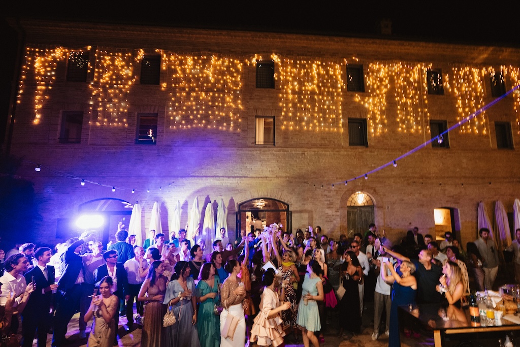 Ospiti in atmosfera serale con edificio rustico addobbato da lucine calde sullo sfondo - ACCAELLE Events | Organizzazione eventi e Wedding planner