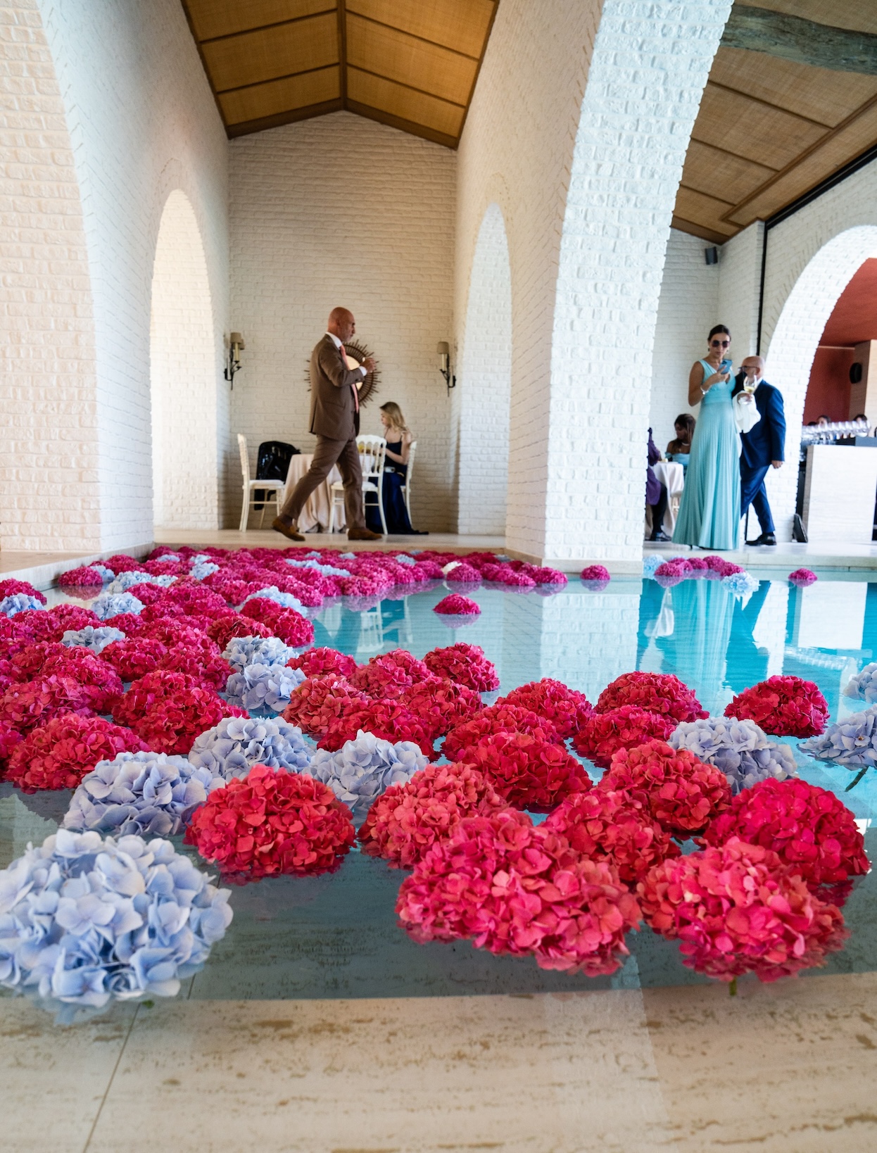 Piscina interna posta in una struttura bianca decorata con fiori in ammollo di colore rosa e lilla e persone sullo sfondo - ACCAELLE Events | Organizzazione eventi e Wedding planner