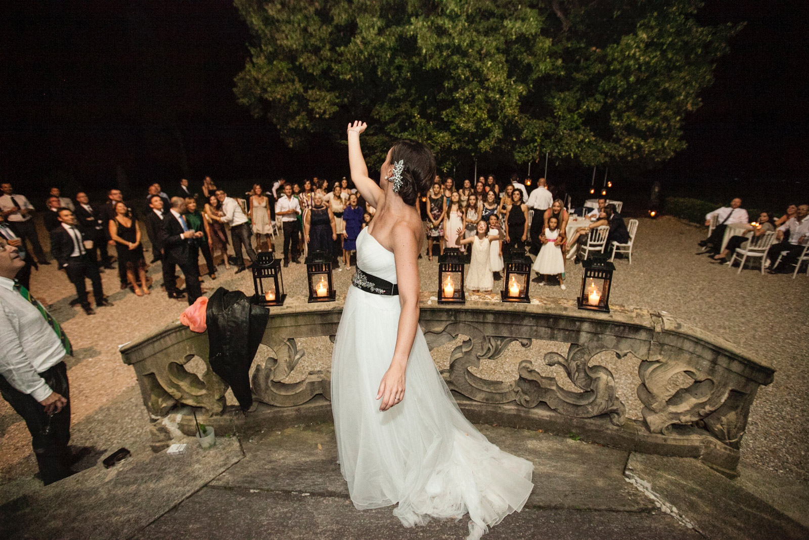 Sposa di spalle con braccia alzato con ospiti sullo sfondo di sera - ACCAELLE Events | Organizzazione eventi e Wedding planner