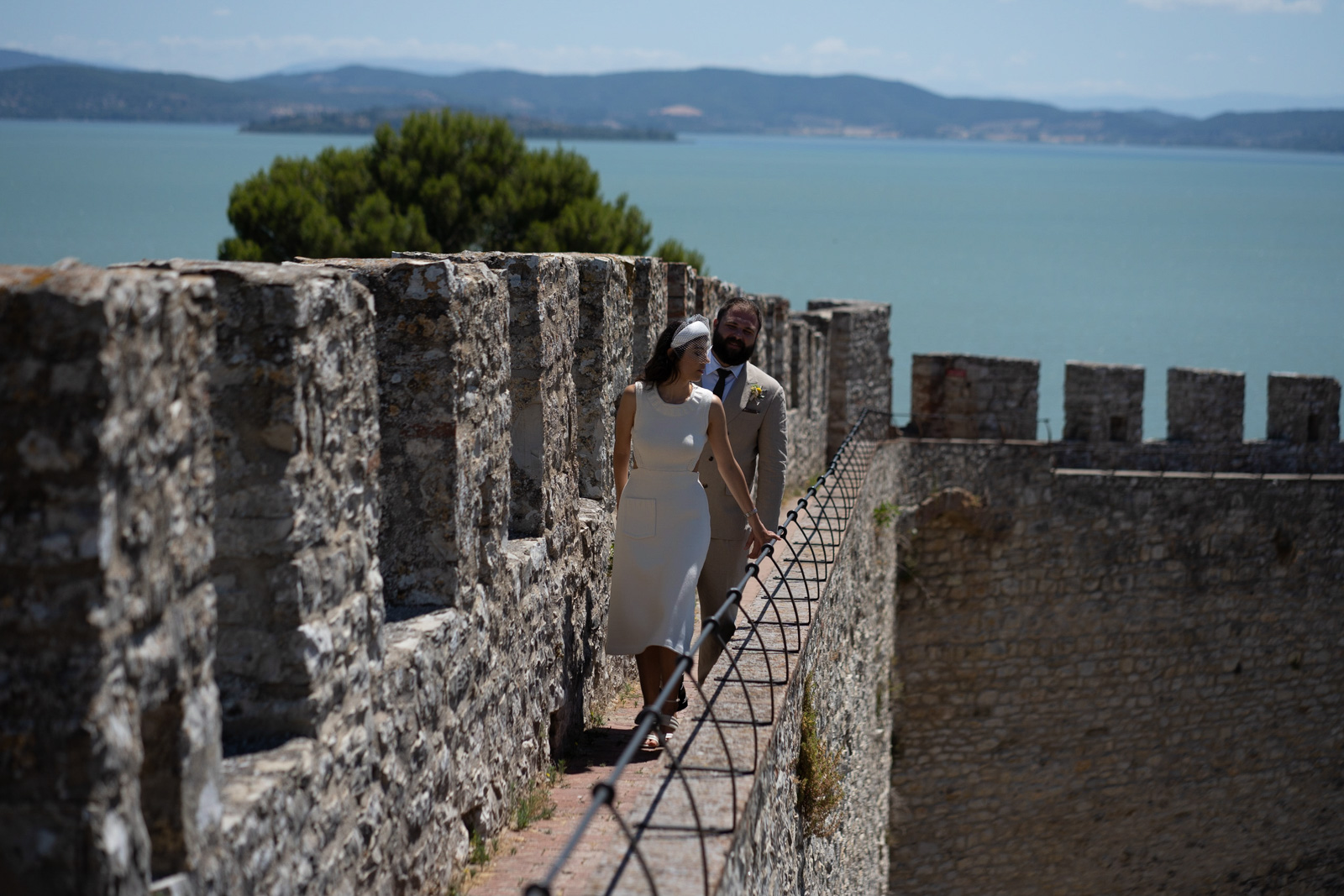 Sposi camminano sulle mura di un castello medievale in pietra, con un grande lago sullo sfondo. - ACCAELLE Events | Organizzazione eventi e Wedding planner