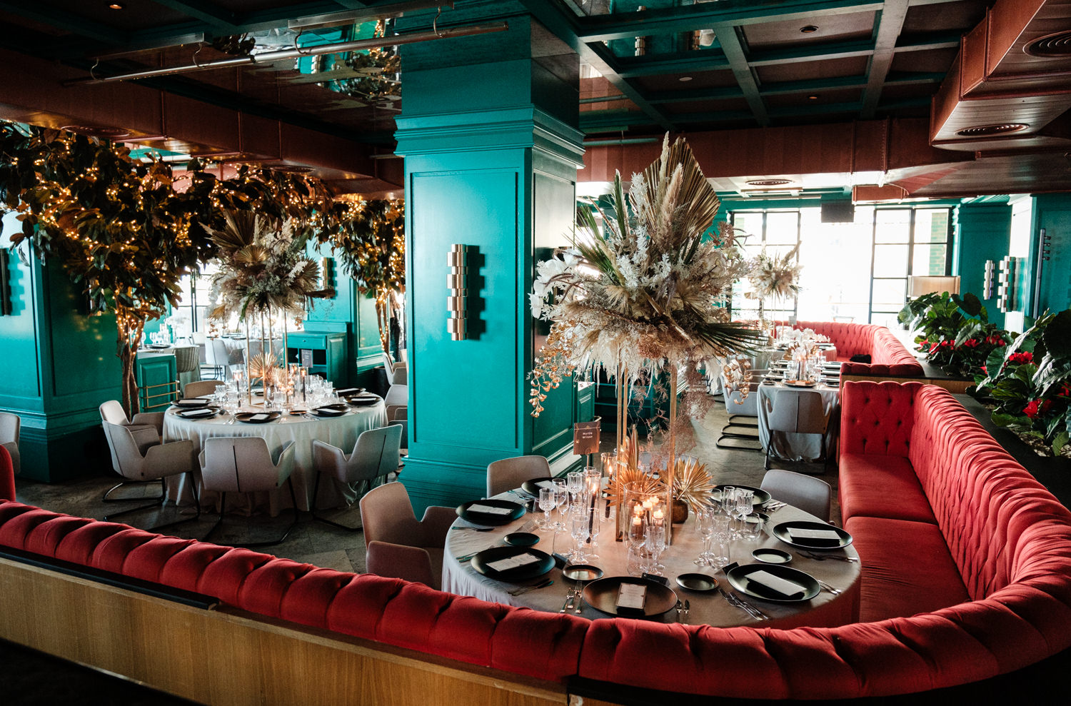 Tavoli rotondi con poltroncine bianche in una sala con pareti color verde acqua, divanetti rossi e composizioni floreali - ACCAELLE Events | Organizzazione eventi e Wedding planner