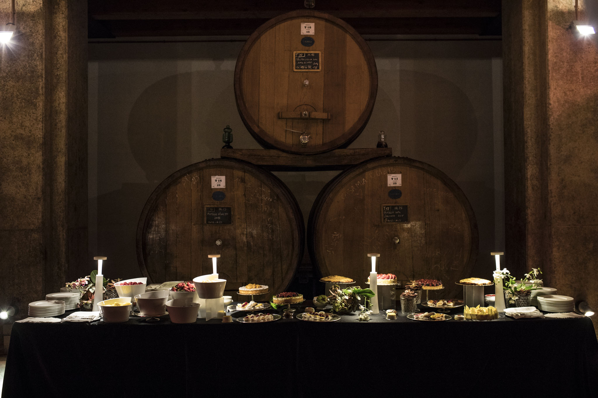Tavolo del buffet allestito davanti a tre grandi botti di legno in una cantina. - ACCAELLE Events | Organizzazione eventi e Wedding planner