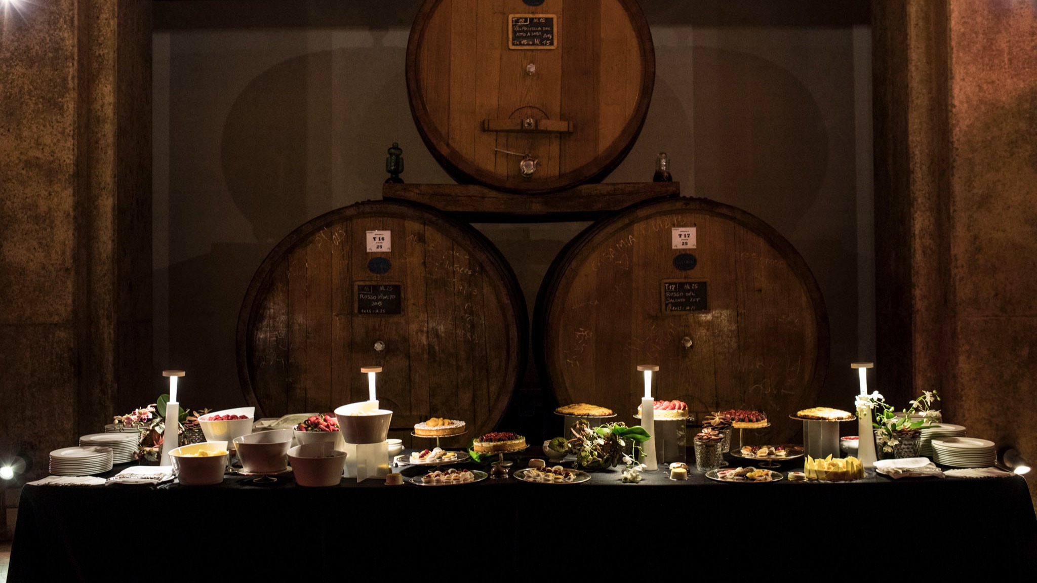Tavolo del buffet allestito davanti a tre grandi botti di legno in una cantina. - ACCAELLE Events | Organizzazione eventi e Wedding planner